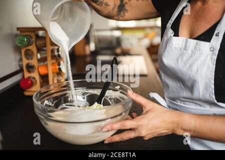 Crop casalinga che indossa grembiule in piedi al banco in cucina e. versare lo zucchero nel recipiente di vetro mentre si prepara l'impasto per la pasta Foto Stock