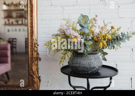 Bel bouquet con vari fiori tra cui il gondrod e fiori di craspedia con rami di eucalipto verde disposti in pentola ornamentale in ceramica su tavolino contro muro bianco di mattoni con specchio in studio di floristry creativo Foto Stock