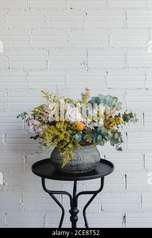 Bel bouquet con vari fiori tra cui il gondrod e fiori di craspedia con rami di eucalipto verde disposti in pentola ornamentale in ceramica su un piccolo tavolo contro muro di mattoni bianchi in floristry creativo studio Foto Stock