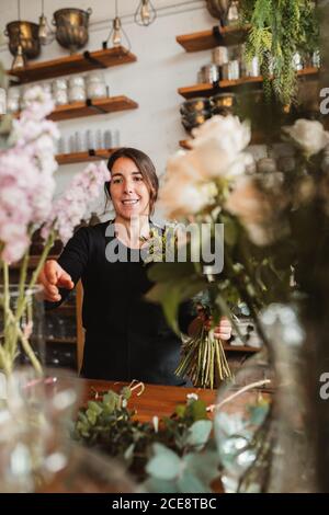 Positivo giovane donna floristry designer con grande bouquet di fresco fiori in fiore e verde fogliame in piedi al bancone in accogliente negozio Foto Stock