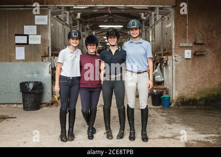 Gruppo di amichevoli equestriani in uniforme abbracciando vicino fienile in campagna mentre si sta insieme e si guarda la fotocamera Foto Stock