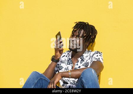 sorridente giovane nero seduto contro una parete gialla che registra un messaggio vocale con il suo smartphone, tecnologia e stile di vita concetto, copia spazio per il testo Foto Stock