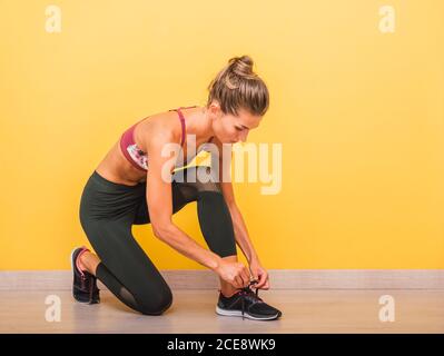 Vista laterale di un sportivo sicuro in reggiseno e leggings che legano le scarpe sulle sneaker prima di allenarsi in palestra moderna Foto Stock