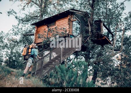 Vista posteriore ad angolo basso del viaggiatore maschile con zaino a piedi su le scale in una casa di cabina su albero Foto Stock