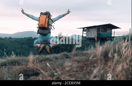 Vista posteriore di un allegro zaino in spalla maschile nel momento del salto con braccia distese sullo sfondo di paesaggio montuoso in serata Foto Stock