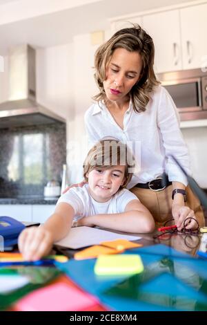 Donna adulto seria in vestito formale revisione dei compiti in piedi vicino sorridente ragazzo seduto a tavola con adesivi multicolore mentre studiano in cucina Foto Stock