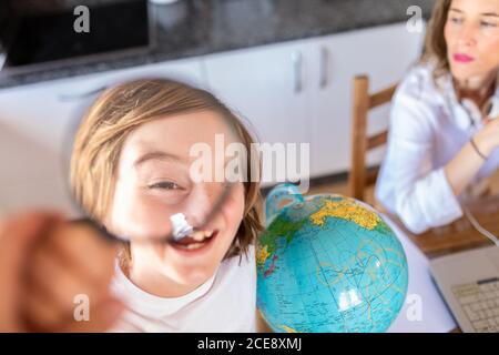 Dalla parte superiore del ragazzo positivo guardando attraverso la lente di ingrandimento mentre in piedi vicino al globo colorato e raccolto premuroso Donna seduto vicino al portatile a tavolo a casa Foto Stock