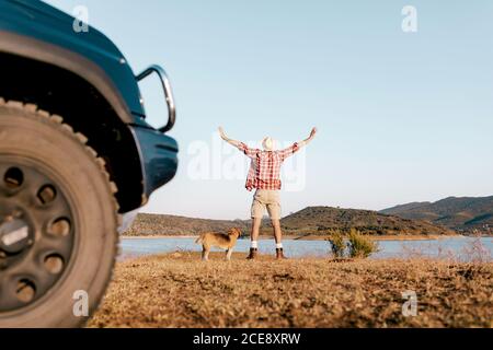 Vista posteriore di turista maschile anonimo in abbigliamento casual e. cappello in piedi su erba sbiadita con le mani a parte vicino Hovawart ammira il lago circondato dai monti durante il viaggio in auto Foto Stock