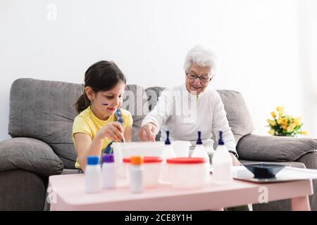 Sorridente erboso in occhiali aiutando nipote positiva con faccia sporca in abbigliamento casual preparazione di una miscela con coloranti in contenitore di plastica mentre si è seduti su un comodo divano nel soggiorno Foto Stock