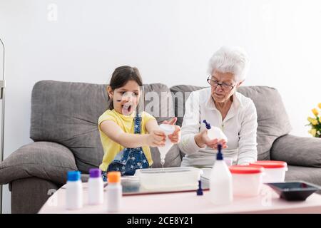 Sorridente erboso in occhiali aiutando nipote positiva con faccia sporca in abbigliamento casual preparazione di una miscela con coloranti in contenitore di plastica mentre si è seduti su un comodo divano nel soggiorno Foto Stock