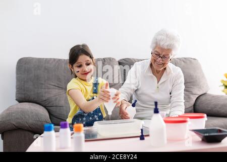 Sorridente erboso in occhiali aiutando nipote positiva con faccia sporca in abbigliamento casual preparazione di una miscela con coloranti in contenitore di plastica mentre si è seduti su un comodo divano nel soggiorno Foto Stock