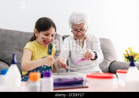 Sorridente erboso in occhiali aiutando nipote positiva con faccia sporca in abbigliamento casual preparazione di una miscela con coloranti in contenitore di plastica mentre si è seduti su un comodo divano nel soggiorno Foto Stock