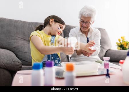 Sorridente erboso in occhiali aiutando nipote positiva con faccia sporca in abbigliamento casual preparazione di una miscela con coloranti in contenitore di plastica mentre si è seduti su un comodo divano nel soggiorno Foto Stock