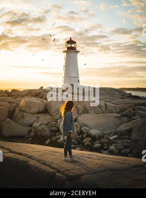 Vista laterale di anonima Donna in piedi su sentiero di pietra e guardando Peggys Cove Lighthouse e uccelli volanti contro cielo nuvoloso tramonto in Canada Foto Stock