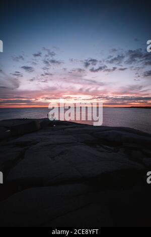 Costa sassosa con sagome di persone situate contro l'acqua di mare e cielo nuvoloso tramonto in serata a Peggys Cove, Canada Foto Stock