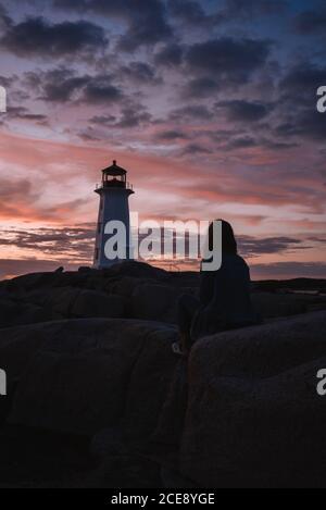 Vista posteriore della donna anonima seduta sul sentiero di pietra e guardando il faro di Peggys Cove contro il cielo nuvoloso del tramonto in Canada Foto Stock