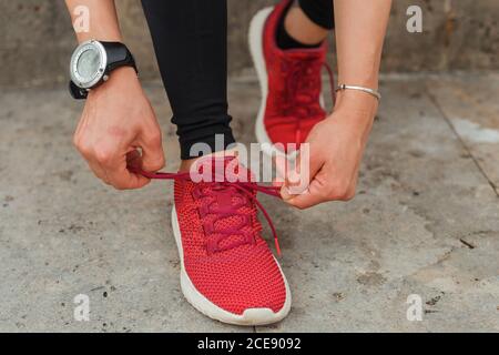 Dall'alto di raccolto anonimo atleta femminile in classico indossabile braccialetto che lega le scarpe rosse mentre sbatte sul marciapiede di luce diurna Foto Stock