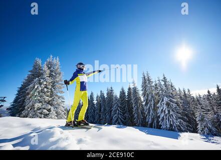 Foto a basso angolo di sciatore femminile prima di sciare da discesa. Donna sorridente in occhiali che mostra con pali da sci su sole luminoso in cielo blu. Alberi di abete innevati sullo sfondo. Stile di vita ricreativo. Foto Stock