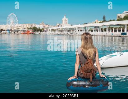 Vista posteriore della donna tatuata con piccolo zaino seduto vicino all'acqua con le mani a parte e guardando la zona costiera della città Foto Stock