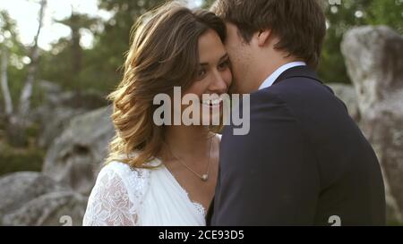 Uomo baciare bella Donna in orecchio mentre sorridendo e carezzandosi felicemente alla luce del sole sulla costa con alberi verdi Foto Stock