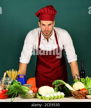 Cucina lavori in cucina vicino a tavola con verdure e utensili. Uomo in burgundy cuoco cappello e grembiule. Chef con viso arrabbiato si appoggia sul tavolo su sfondo verde scuro. Concetto di pasto vegetariano. Foto Stock