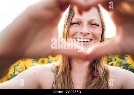 Primo piano ritratto di giovane donna sorridente che fa la forma del cuore con le mani al campo di girasole Foto Stock