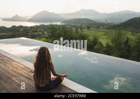 Donna che medita e pratica yoga da sola all'alba vicino piscina infinity con montagne all'orizzonte. Vista posteriore. Concetto di rilassamento spirituale stile di vita di viaggio. Armonia con la natura. Foto Stock