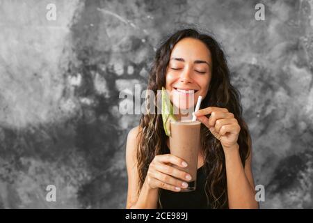 Divertente donna felice bere latte di cioccolato agitare con cacao e ridere. Concetto di bevanda estiva. Fondo parete in cemento grigio. Foto Stock