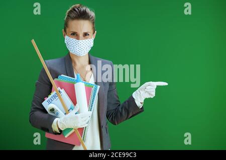 moderna insegnante di scuola femminile in blusa bianca con maschera medica, guanti di gomma, termometro digitale, libro di testo e igienizzatore che indica qualcosa sulla cha Foto Stock