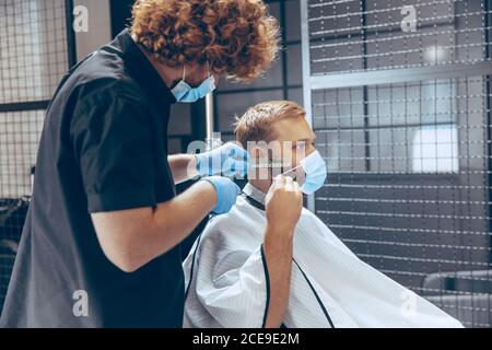 Uomo che ottiene i capelli tagliati al barbiere indossando la maschera durante la pandemia del coronavirus. Guanti professionali da barbiere. Covid-19, concetto di bellezza, autosocura, stile, salute e medicina. Foto Stock