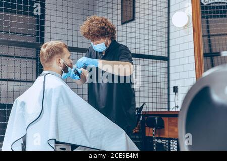 Uomo che ottiene i capelli tagliati al barbiere indossando la maschera durante la pandemia del coronavirus. Guanti professionali da barbiere. Covid-19, concetto di bellezza, autosocura, stile, salute e medicina. Foto Stock