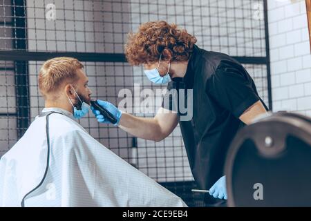 Uomo che ottiene i capelli tagliati al barbiere indossando la maschera durante la pandemia del coronavirus. Guanti professionali da barbiere. Covid-19, concetto di bellezza, autosocura, stile, salute e medicina. Foto Stock