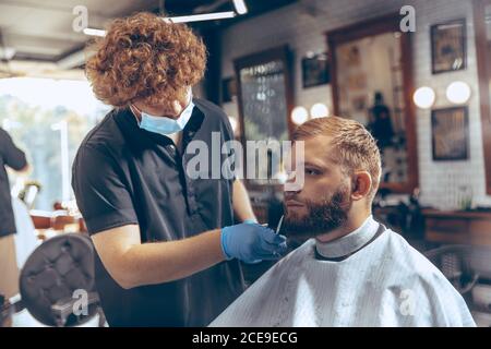 Uomo che ottiene i capelli tagliati al barbiere indossando la maschera durante la pandemia del coronavirus. Guanti professionali da barbiere. Covid-19, concetto di bellezza, autosocura, stile, salute e medicina. Foto Stock