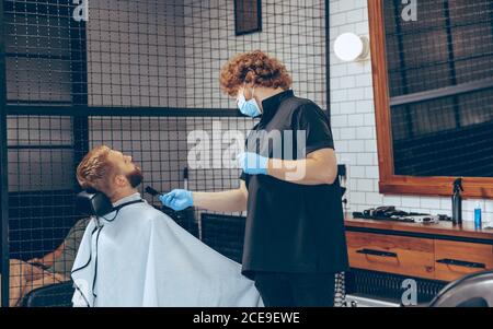 Uomo che ottiene i capelli tagliati al barbiere indossando la maschera durante la pandemia del coronavirus. Guanti professionali da barbiere. Covid-19, concetto di bellezza, autosocura, stile, salute e medicina. Foto Stock