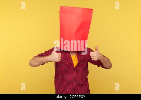 Ragazzo in camicia a scacchi in piedi con la borsa della spesa sulla testa, mostrando pollici come gesto, consigliando negozio di moda, vendite e sconti. Indoor Foto Stock