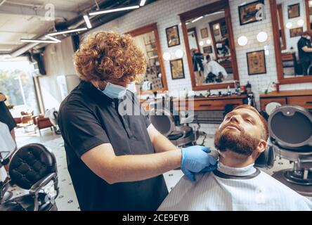 Uomo che ottiene i capelli tagliati al barbiere indossando la maschera durante la pandemia del coronavirus. Guanti professionali da barbiere. Covid-19, concetto di bellezza, autosocura, stile, salute e medicina. Foto Stock