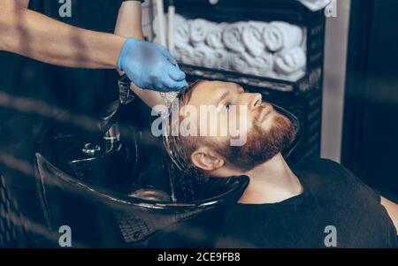 Uomo che ottiene i capelli tagliati al barbiere indossando la maschera durante la pandemia del coronavirus. Guanti professionali da barbiere. Covid-19, concetto di bellezza, autosocura, stile, salute e medicina. Foto Stock