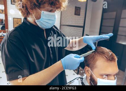 Uomo che ottiene i capelli tagliati al barbiere indossando la maschera durante la pandemia del coronavirus. Guanti professionali da barbiere. Covid-19, concetto di bellezza, autosocura, stile, salute e medicina. Foto Stock