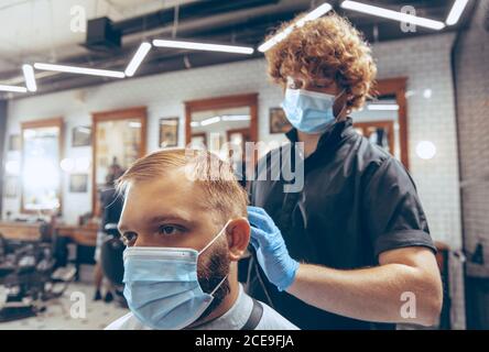 Uomo che ottiene i capelli tagliati al barbiere indossando la maschera durante la pandemia del coronavirus. Guanti professionali da barbiere. Covid-19, concetto di bellezza, autosocura, stile, salute e medicina. Foto Stock