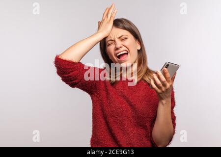 Ritratto di donna depressa sconvolta in maglione shaggy in piedi con gesto facepalm e tenendo il telefono cellulare, esprimendo dolore rammarico, incolpando se stessa Foto Stock