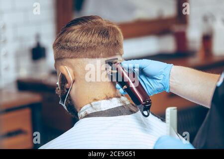 Uomo che ottiene i capelli tagliati al barbiere indossando la maschera durante la pandemia del coronavirus. Guanti professionali da barbiere. Covid-19, concetto di bellezza, autosocura, stile, salute e medicina. Foto Stock