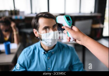 Uomo d'affari con maschera facciale che lavora all'interno in ufficio, misurando la temperatura. Foto Stock