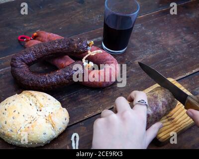 Una persona che taglia una salsiccia di maiale iberica su un vecchio tavola di legno con un coltello antico Foto Stock