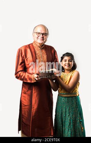 L'anziano o l'anziano indiano con la nipote che festeggia il compleanno soffiando le candele sulla torta mentre indossa l'usura etnica, in piedi isolato contro il bianco Foto Stock