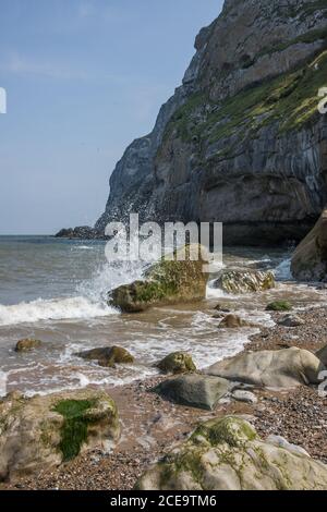 Onde spruzzi sulle rocce alla base del piccolo Orme, Llandudno. North Wales spiaggia di ciottoli Foto Stock