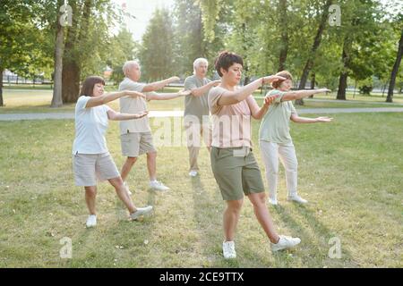 Gruppo di persone senior focalizzate e la loro allenatore gesturing mani e fare esercizio di qigong nel parco Foto Stock