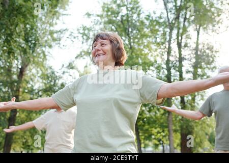 Sotto la vista di allegra donna anziana energica in tshirt outstretching armi mentre si gode l'esercizio mattutino nel parco Foto Stock