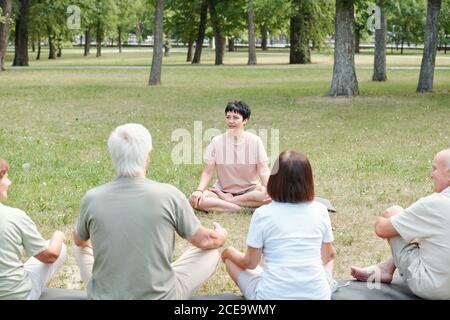 Allenatore professionale di yoga seduto in posizione lotus nel parco e. spiegare le basi dello yoga agli studenti più anziani Foto Stock