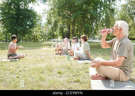 Uomo anziano con baffi seduti con gambe incrociate sul tappetino nel parco e nell'acqua potabile dopo la pratica di yoga Foto Stock