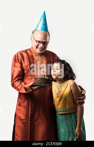 L'anziano o l'anziano indiano con la nipote che festeggia il compleanno soffiando le candele sulla torta mentre indossa l'usura etnica, in piedi isolato contro il bianco Foto Stock